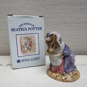 May include: A ceramic figurine of a rabbit wearing a purple coat, white apron, and bonnet, standing on a stone-textured base. Beside it is a light blue cardboard box labelled THE WORLD OF BEATRIX POTTER and ROYAL ALBERT with a printed illustration.