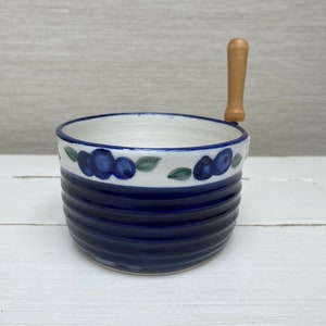 Puede incluir: Un cuenco de cerámica azul y blanco con un asa de madera. El cuenco tiene un diseño de arándanos y es perfecto para servir aperitivos o salsas.