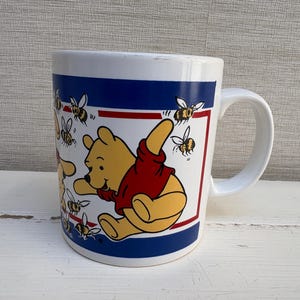 Peut inclure: Un mug en céramique blanche orné d'une illustration de Winnie l'ourson portant un t-shirt rouge, entouré de plusieurs petites abeilles noires et jaunes. Le motif est encadré par des bandes horizontales bleues et rouges sur le pourtour.