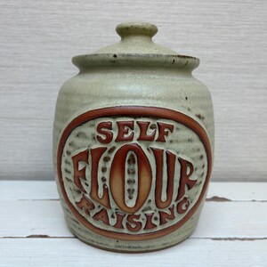 May include: A ceramic jar with a lid, featuring the words "SELF FLOUR RAISING" in a brown oval. The jar has a light green color with a textured surface. The lid has a small knob.
