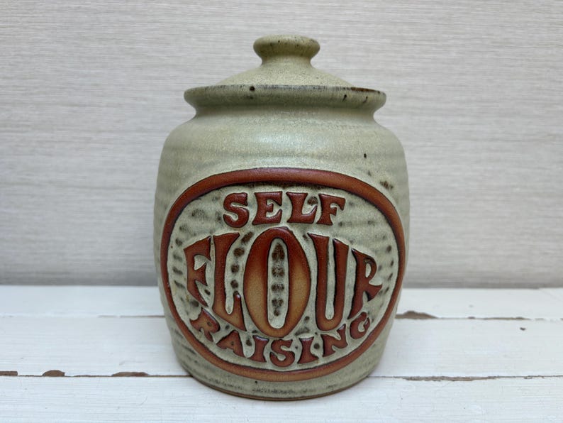 May include: A ceramic jar with a lid, featuring the words "SELF FLOUR RAISING" in a brown oval. The jar has a light green color with a textured surface. The lid has a small knob.
