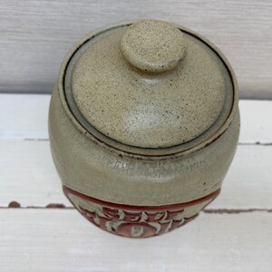 May include: A round, lidded ceramic jar with a speckled, light tan finish. The jar has a reddish-brown band with embossed text near the base. The lid has a small, round knob. The jar is likely for kitchen or home decor.