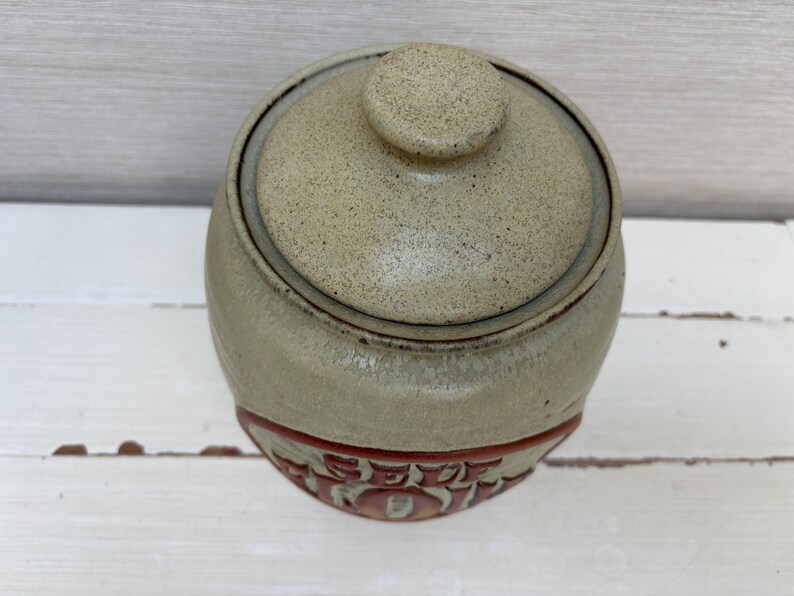May include: A round, lidded ceramic jar with a speckled, light tan finish. The jar has a reddish-brown band with embossed text near the base. The lid has a small, round knob. The jar is likely for kitchen or home decor.
