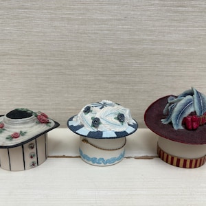 Könnte beinhalten: Drei dekorative hutförmige Boxen mit Blumen- und Federmotiven. Die Boxen sind weiß mit schwarzen, blauen und roten Akzenten.