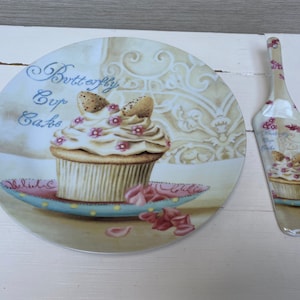 Könnte beinhalten: Ein weißer Keramik-Kuchenteller mit einem Schmetterlings-Cupcake-Design. Der Teller zeigt einen Cupcake mit rosa Zuckerguss und einem Schmetterling aus Schokolade. Der Text "Butterfly Cup Cakes" ist auf dem Teller geschrieben. Ein passender weißer Keramik-Kuchenschieber mit einem Schmetterlings-Cupcake-Design ist ebenfalls enthalten.