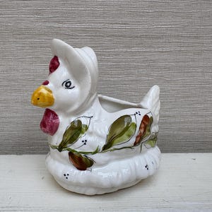 Könnte beinhalten: Weißer Keramik-Pflanztopf in Form eines Hahns mit grünen, braunen und roten Maldetails. Der Topf hat eine kleine Öffnung oben zum Pflanzen.