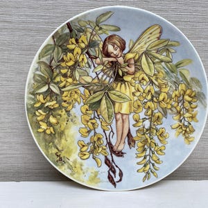 Könnte beinhalten: Ein Porzellanteller mit einem zarten Feendesign. Die Fee ist mit Flügeln und einem gelben Kleid dargestellt, eingebettet in gelbe Blumen und grüne Blätter. Der Teller hat einen hellblauen Hintergrund.