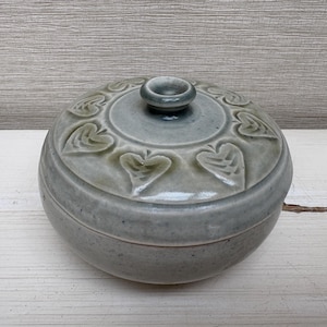 Könnte beinhalten: Runde, graue Keramikdose mit Deckel und matter Oberfläche. Der Deckel hat einen zentralen Knopf und ist mit geprägten herzförmigen Blattmotiven verziert. Ein dekorativer Aufbewahrungsbehälter.