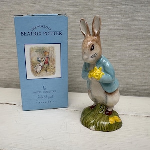 May include: A ceramic figurine of a brown rabbit wearing a blue jacket and holding yellow flowers, standing on a green base. Beside it is a light blue cardboard box labelled The World of Beatrix Potter and Royal Doulton Studios.