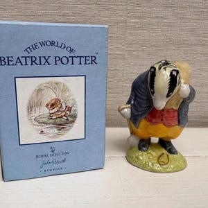 Może przedstawiać: Figurka Royal Doulton przedstawiająca borsuka w niebieskiej kurtce, czerwonej kamizelce i żółtych spodniach, niosącego worek. Figurka stoi na zielonej podstawie. Obok niebieskie pudełko z napisem "The World of Beatrix Potter" i ilustracją.