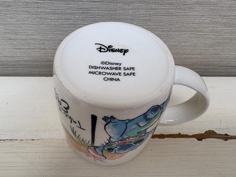 May include: White ceramic mug featuring a colorful illustration of Stitch from Disney's Lilo & Stitch. The mug has a curved handle and the text "Take Care" is visible. The bottom of the mug has the Disney logo and safety information.