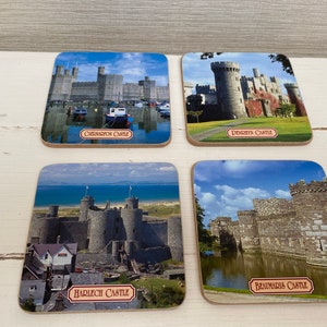 Könnte beinhalten: Vier quadratische Untersetzer mit Bildern verschiedener Burgen in Wales. Die Untersetzer sind aus Kork und haben einen braunen Rand. Die Burgen sind vor blauem Himmel und grünem Gras dargestellt. Die Untersetzer sind mit den Namen der Burgen beschriftet: Caernarfon Castle, Denbigh Castle, Harlech Castle und Beaumaris Castle.