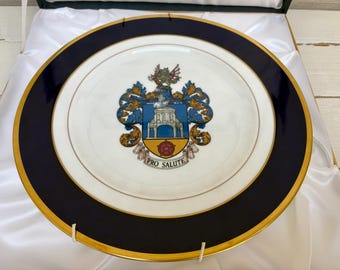 May include: A decorative plate with a navy blue and gold rim. The center features a detailed crest with a building, a rose, and the words "PRO SALUTE". The plate is presented in a green and white box.