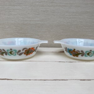 May include: Two white ceramic bowls with floral designs in shades of brown, green, and blue. The bowls have handles on either side.