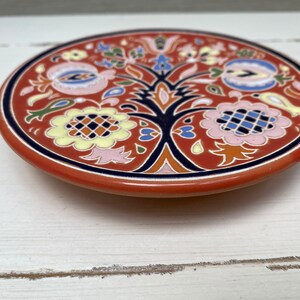 May include: A round ceramic coaster with a red background and a floral design in blue, yellow, pink, and green. The design features a stylised tree with flowers and leaves.