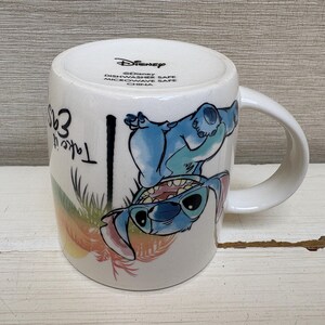 May include: White ceramic mug featuring a blue cartoon character with sunglasses. The mug has a curved handle and the words "Take it easy" are printed on the side. The mug is a Disney product.