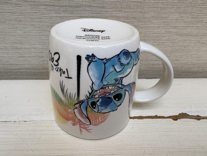 May include: White ceramic mug featuring a blue cartoon character with sunglasses. The mug has a curved handle and the words "Take it easy" are printed on the side. The mug is a Disney product.