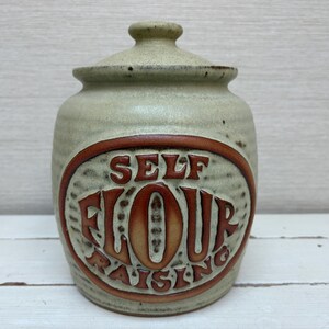May include: A ceramic jar with a lid, featuring the words "SELF FLOUR RAISING" in a brown oval. The jar is a light green color with a brown rim and lid, suitable for kitchen storage.