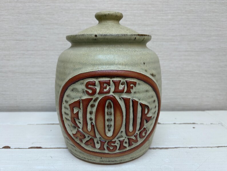 May include: A ceramic jar with a lid, featuring the words "SELF FLOUR RAISING" in a brown oval. The jar is a light green color with a brown rim and lid, suitable for kitchen storage.