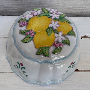May include: White ceramic serving dish with a lemon and floral design. The lid features three yellow lemons, green leaves, and pink and white flowers. The rim has a light blue trim and decorative green and maroon accents.
