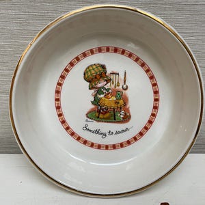 Könnte beinhalten: Eine weiße Keramikschüssel mit goldfarbenem Rand und einem rot-weiß karierten Muster. Die Schüssel zeigt eine Comic-Illustration eines Mädchens beim Backen mit dem Text "Something to savour..."