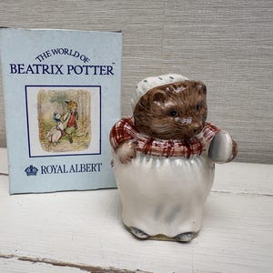 Peut inclure: Une figurine en céramique représentant un hérisson brun portant un bonnet blanc et un châle à carreaux rouges et blancs, à côté d'une boîte bleu clair marquée The World of Beatrix Potter et Royal Albert avec une illustration d'un canard et d'un renard.