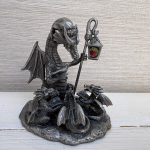 Könnte beinhalten: Eine silberfarbene Metallfigur, die einen großen Drachen zeigt, der einen Stab mit einer hängenden Laterne hält, in der ein buntes Licht leuchtet. Zwei kleinere Drachen sitzen an der Basis und halten jeweils ein kleines Buch auf einem strukturierten Sockel.