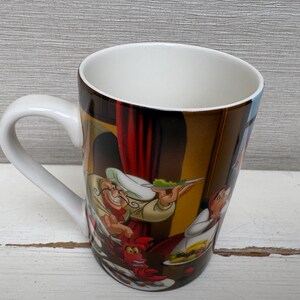May include: White ceramic coffee mug with a curved handle. The mug features a colorful illustration of cartoon characters in a kitchen scene. The interior of the mug is white.