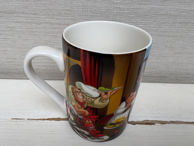 May include: White ceramic coffee mug with a curved handle. The mug features a colorful illustration of cartoon characters in a kitchen scene. The interior of the mug is white.
