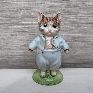 May include: A ceramic figurine of a brown tabby kitten standing upright. The kitten wears a light blue jacket and matching trousers with small gold-colored buttons. It is positioned on a circular green base that resembles a patch of grass.