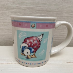 Puede incluir: Una taza de cerámica blanca con un diseño azul, rosa y verde que presenta un pato de dibujos animados. El pato lleva una camisa con estampado rojo y blanco y está mirando hacia abajo a un objeto pequeño.