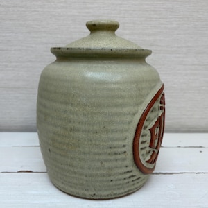 May include: A ceramic jar with a lid, in a muted green color. The jar has a cylindrical shape with a textured surface. A circular design with a reddish-brown border and a carved pattern is on the side. The lid has a small knob.