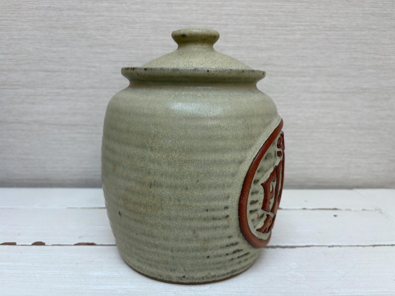 May include: A ceramic jar with a lid, in a muted green color. The jar has a cylindrical shape with a textured surface. A circular design with a reddish-brown border and a carved pattern is on the side. The lid has a small knob.