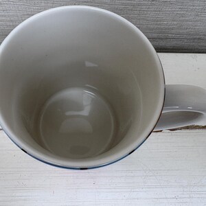 May include: A white ceramic mug with a rounded handle. The interior is white, and the rim has a thin blue and red border. The mug is sitting on a white wooden surface.