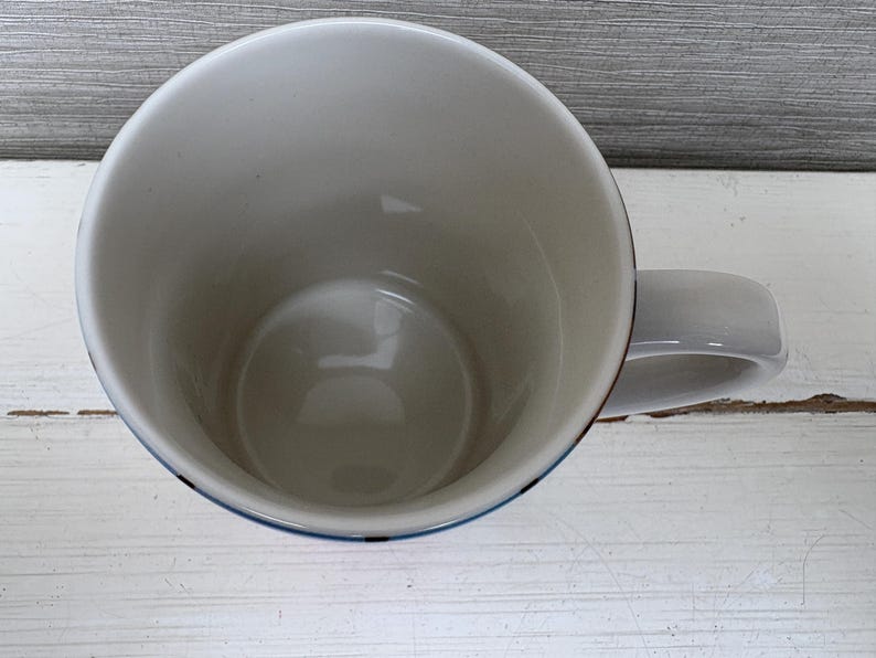 May include: A white ceramic mug with a rounded handle. The interior is white, and the rim has a thin blue and red border. The mug is sitting on a white wooden surface.