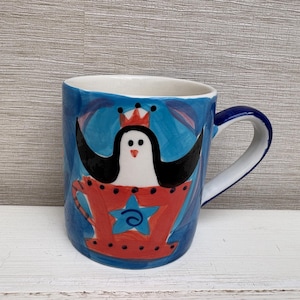 Könnte beinhalten: Handbemalte Keramiktasse mit einem skurrilen Design. Die Tasse hat einen blauen Hintergrund mit einem Pinguin mit Krone in einer roten Tasse. Der Griff ist blau und das Innere ist weiß.