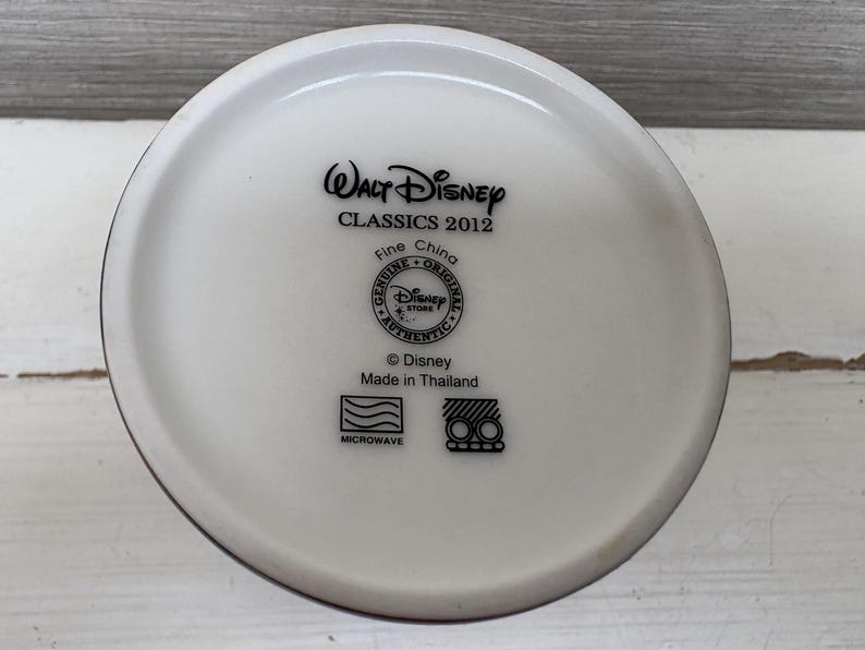 May include: A white ceramic mug base with black text that reads "Walt Disney CLASSICS 2012 Fine China Genuine Original Disney Store Authentic &copy; Disney Made in Thailand Microwave".