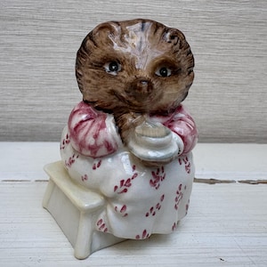 May include: A ceramic hedgehog figurine seated on a small bench. The hedgehog has brown fur and is wearing a white dress with a red floral pattern and a pink shawl. It is holding a small, white object.