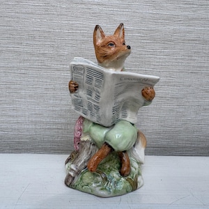 May include: A ceramic figurine of a fox with orange and white fur, wearing light green trousers, sitting on a tree stump whilst holding and reading a white newspaper with black printed text. The base is painted with green and brown details.