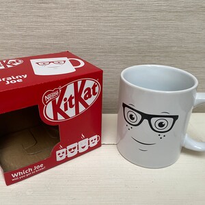 May include: A white ceramic mug with a black and white illustration of a face wearing glasses. The mug is sitting on a white surface. The mug is packaged in a red box with the words "Nestle KitKat" and "Brainy Joe" printed on it.