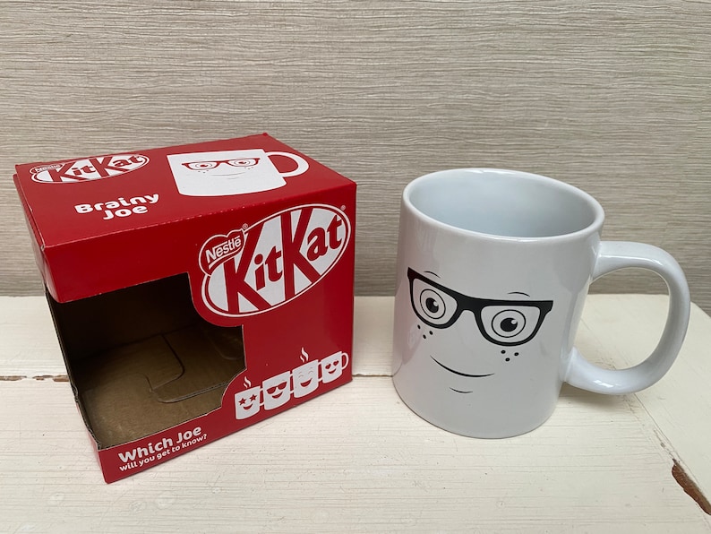 May include: A white ceramic mug with a black and white illustration of a face wearing glasses. The mug is sitting on a white surface. The mug is packaged in a red box with the words "Nestle KitKat" and "Brainy Joe" printed on it.
