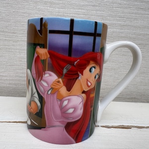May include: A ceramic mug featuring a colorful illustration of a cartoon character with red hair and a pink dress. The mug has a white handle and a blue and brown background. The mug is likely a collectible item.