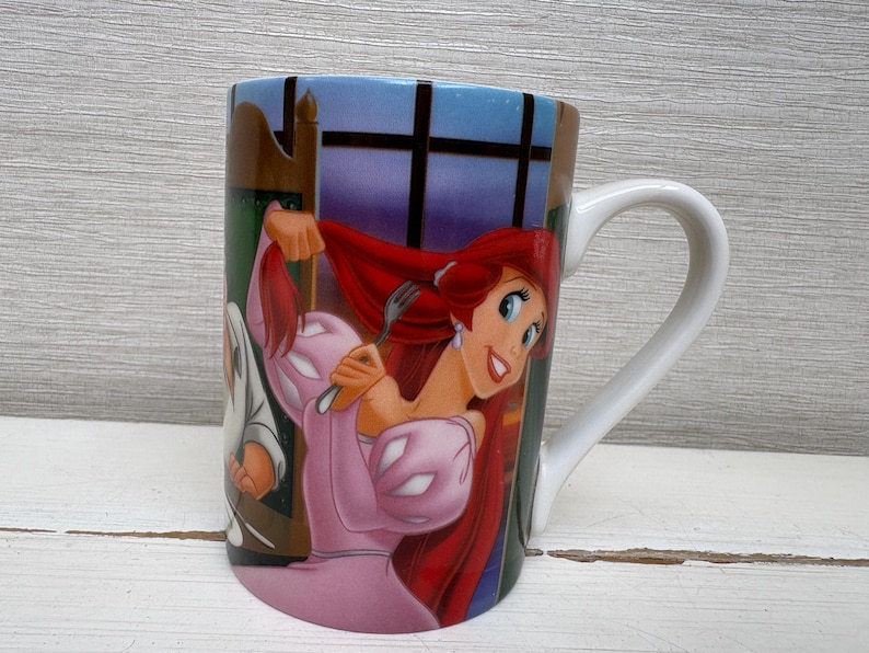 May include: A ceramic mug featuring a colorful illustration of a cartoon character with red hair and a pink dress. The mug has a white handle and a blue and brown background. The mug is likely a collectible item.