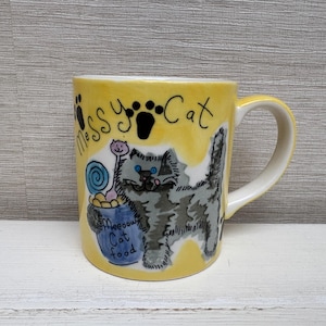 Puede incluir: Taza de cerámica amarilla con una huella de pata negra y las palabras "Messy Cat" en la parte delantera. La taza presenta una ilustración de dibujos animados de un gato gris con un collar azul y un caracol rosa en la espalda. El gato sostiene un tazón azul con las palabras "Meow Cat Food" en él.