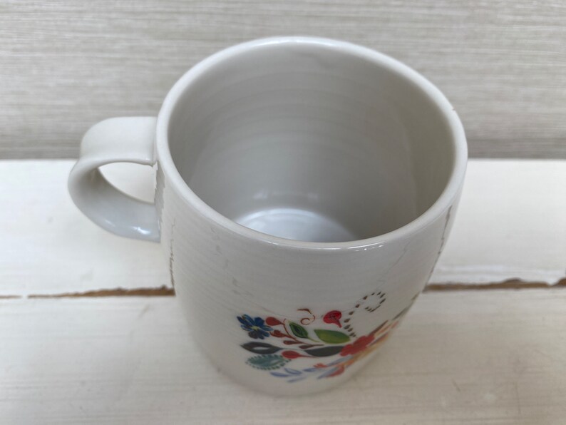 以下が含まれることがあります： 青、赤、緑、黄色の花柄が描かれた白いセラミック製のマグカップ。持ち手と広い縁があります。