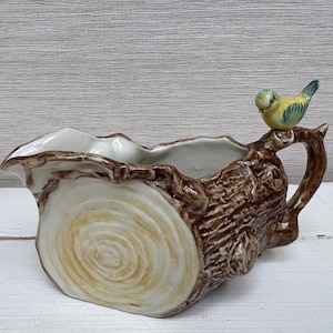 Puede incluir: Una cremera de cerámica con forma de tronco de árbol con un pequeño pájaro posado en el asa. La cremera es de color marrón y crema, con una textura detallada de vetas de madera y un pájaro amarillo, azul y verde.