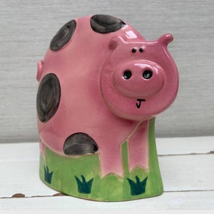 Könnte beinhalten: Eine rosa Keramikschwein-Figur mit schwarzen Flecken und einem grünen Sockel mit Grasdetails. Das Schwein hat ein lächelndes Gesicht und ist ein dekoratives Objekt.