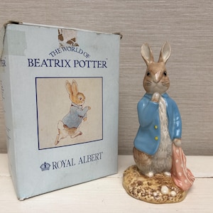 May include: A ceramic Peter Rabbit figurine wearing a blue jacket and holding a pink cloth, standing on a textured base. Beside it is a light blue cardboard box labelled The World of Beatrix Potter and Royal Albert with an illustration of the rabbit.