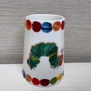 May include: White ceramic tumbler featuring a colorful caterpillar design. The tumbler has a ring of colorful dots around the top and bottom. The caterpillar is green, yellow, and blue with a red head and is a children's theme.