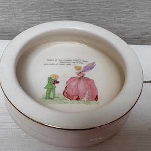 May include: A circular, cream-coloured ceramic bowl with a gold trim. The interior has a colourful illustration of a fairy and a young man, with text above. The bowl has a slightly cracked appearance.